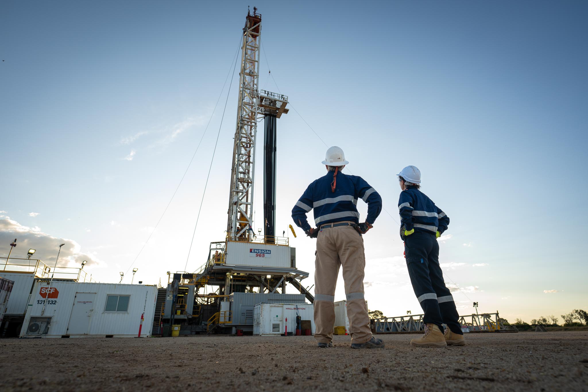 Two workers in hard hats and high visibility workwear stand at a drilling site looking up at a tall rig. The low angle makes the rig appear large against the open sky and shows the scale of the industrial operation.