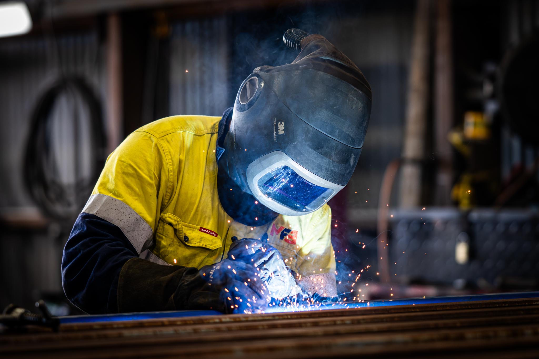 MFE worker welding metal inside a workshop while wearing a protective helmet and high visibility work shirt. Bright sparks and blue welding light show the hands on manufacturing work being captured.