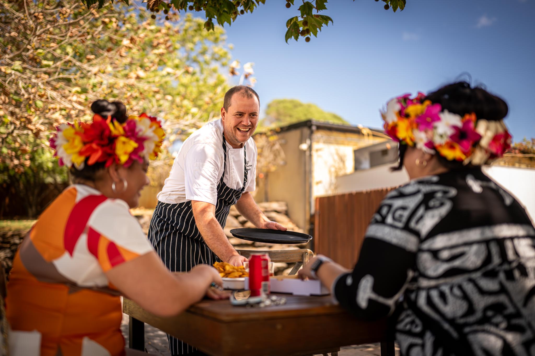 A smiling chef in a white jacket and striped apron serves food to two guests seated at an outdoor table. The guests wear bright flower crowns and the sunny setting gives the photo a warm hospitality feel.