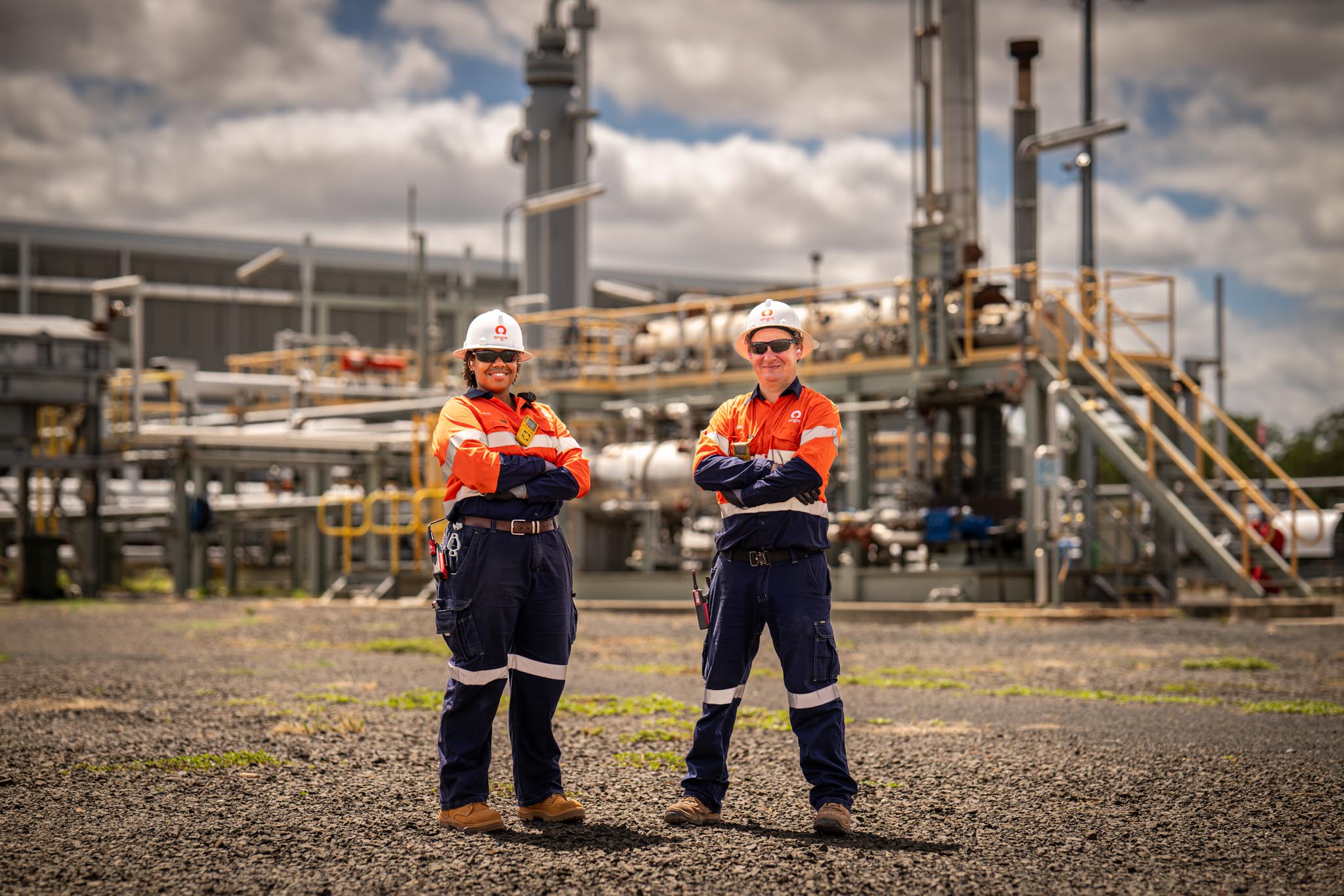 Two Origin Energy workers stand with arms crossed in front of an industrial facility wearing high visibility uniforms and white hard hats. Processing equipment, pipes and metal structures fill the background under a cloudy sky.