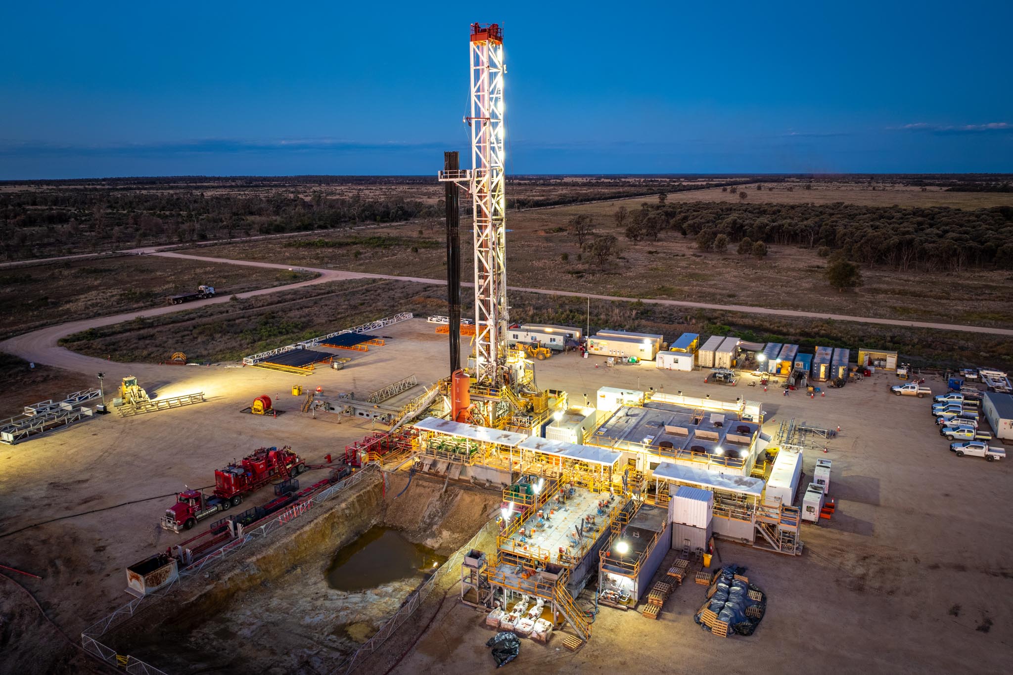 A brightly lit drilling rig stands over a remote worksite at dusk with trucks, site buildings and equipment arranged around it. The surrounding bushland and open roads show the scale of the operation in a rural location.