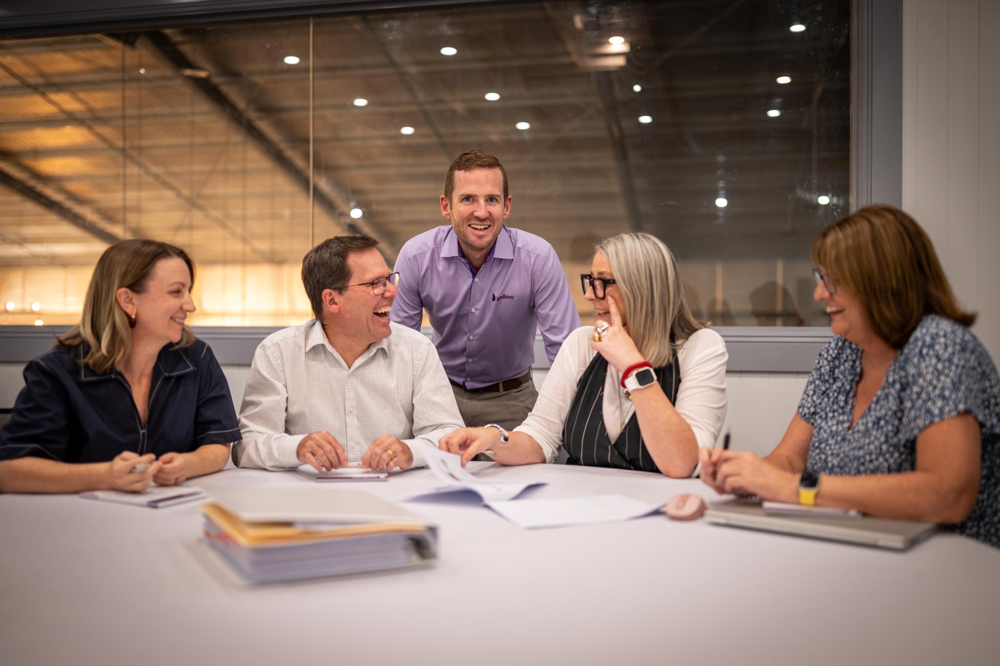 A group of five coworkers sit around a meeting table laughing together with notebooks, papers and folders in front of them. The relaxed office setting shows a collaborative business team during a friendly workplace discussion.