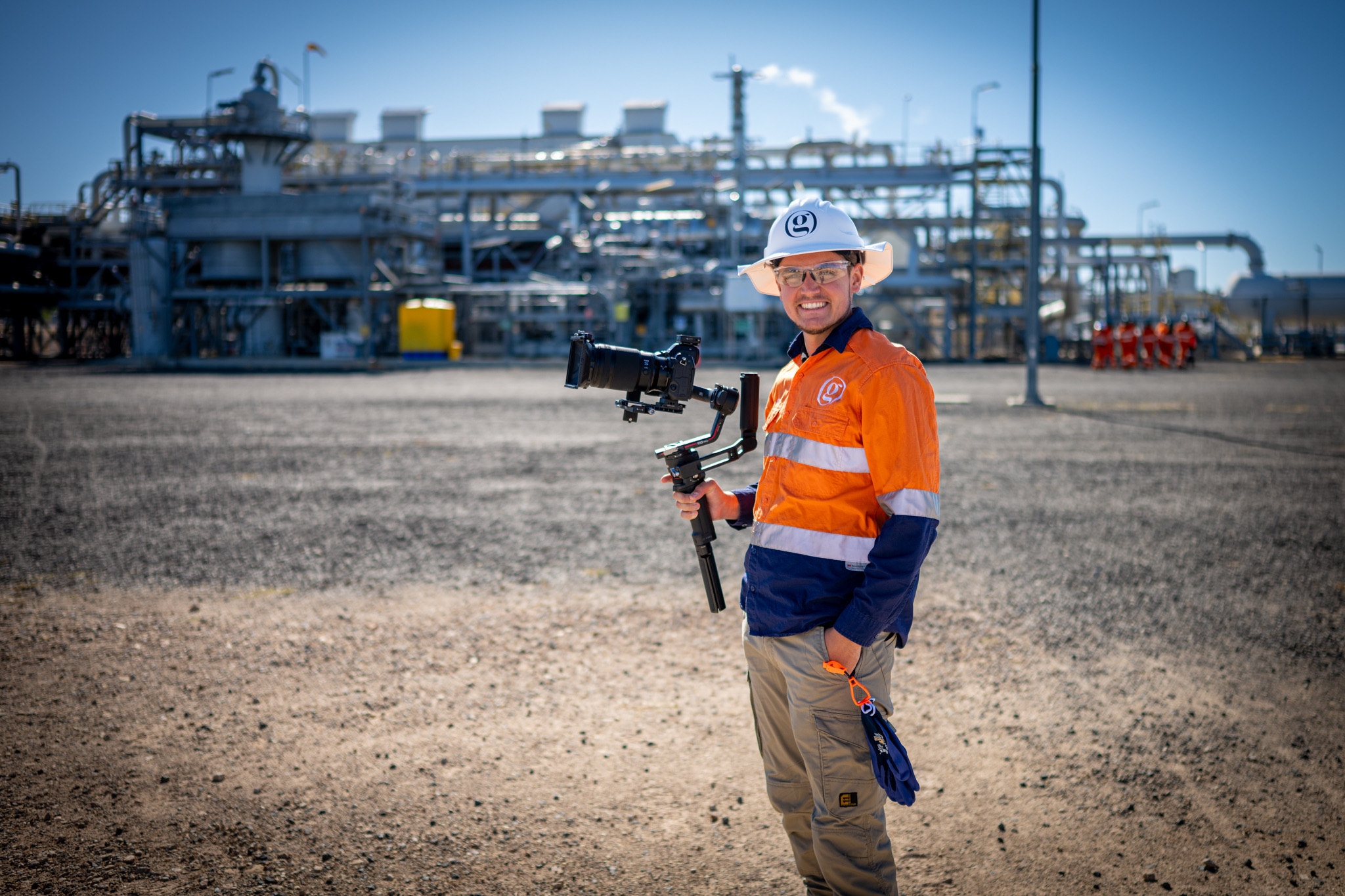 Seth Gerke from Gerkies stands at an industrial site wearing high visibility workwear and a hard hat while holding a camera on a stabiliser. Pipes, processing equipment and workers in the background show Gerkies capturing professional video content in a live work environment.